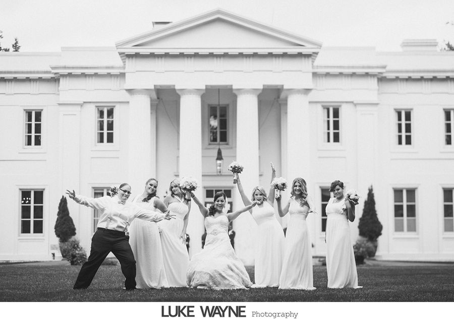 Bride and bridesmaids pose in front of a white building, arms raised and smiling.