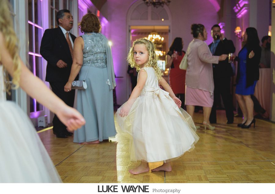A young girl in a blush dress dances barefoot on a wooden dance floor at a party, with other guests in the background.