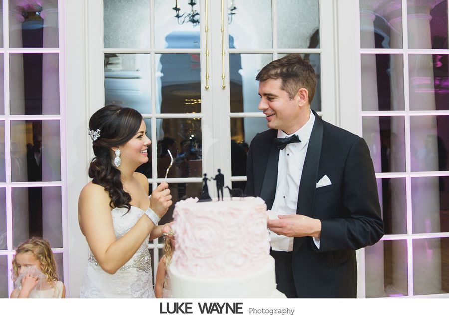 Wedding couple cutting cake; bride smiling, groom looking; pink and white cake, outdoor background.