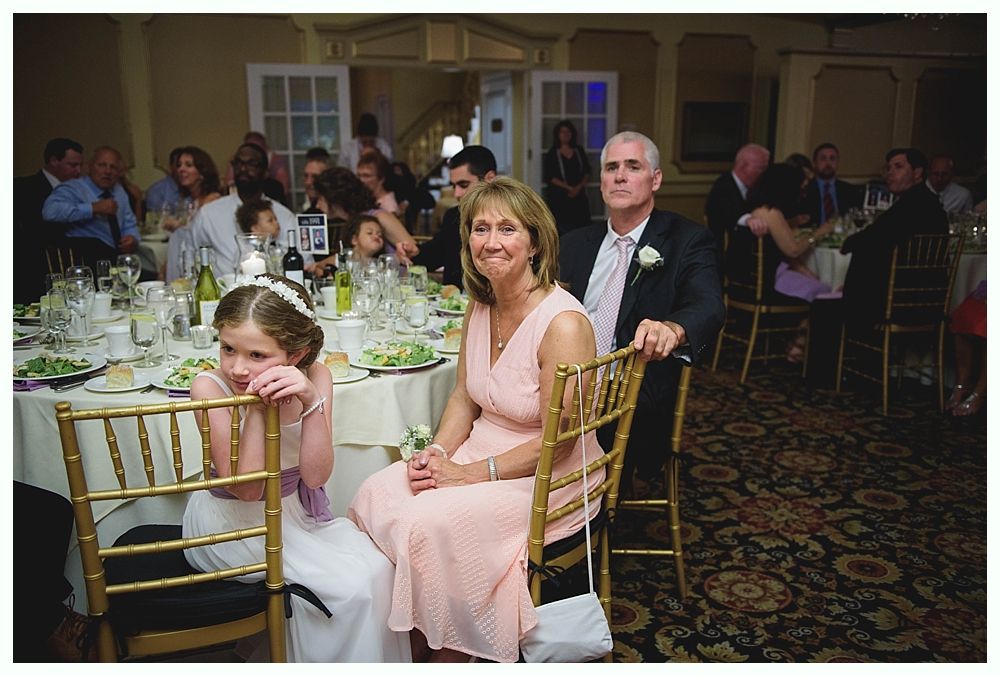 Bride with arms up, making a funny face, groom looking at his hand, cake cutting in background.