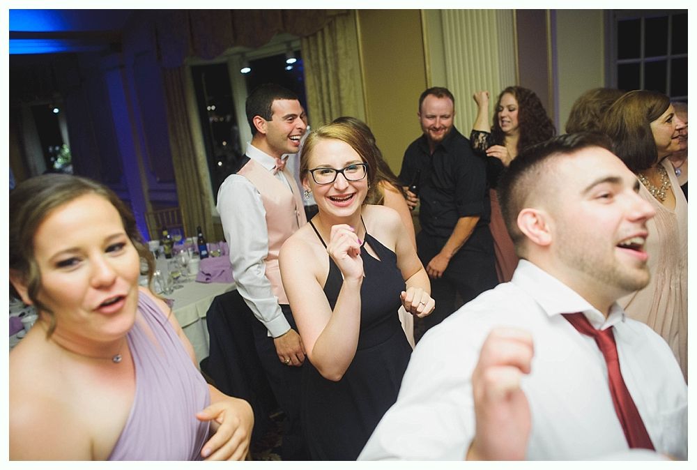Bride with arms up, making a funny face, groom looking at his hand, cake cutting in background.