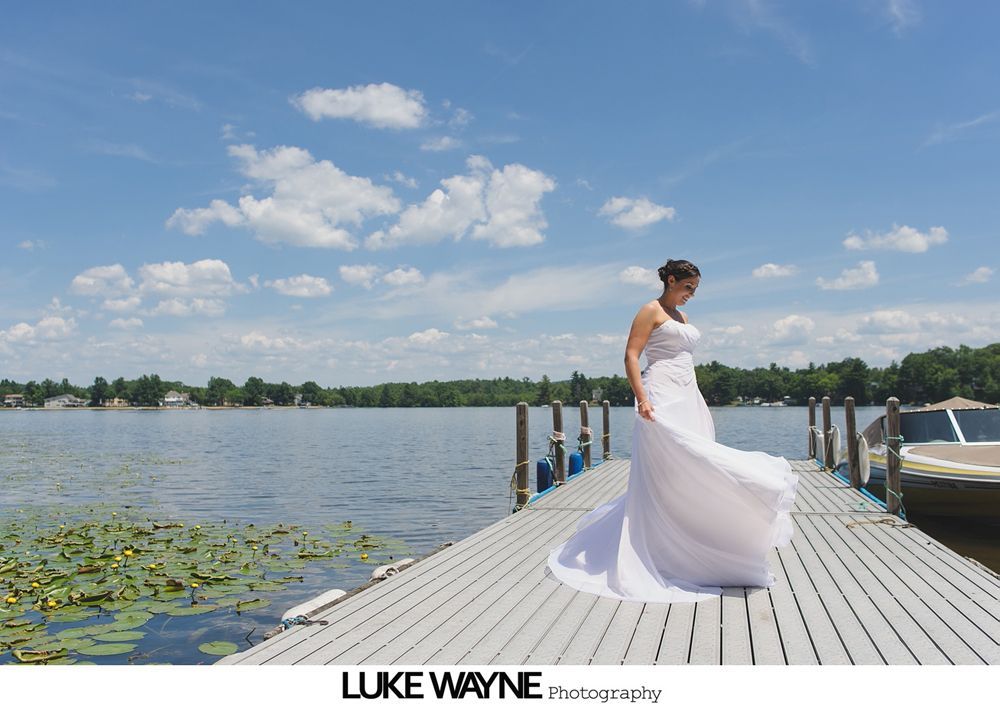 Bride in a white dress on a dock, lake and sky in background.