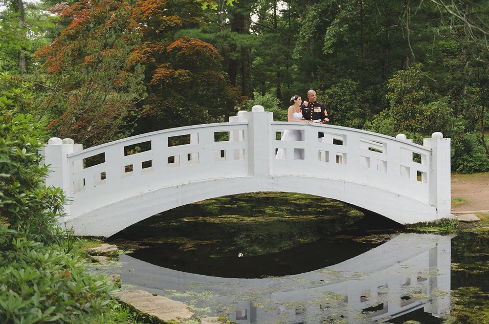 Couple on a white arched bridge over water; lush green trees in the background.