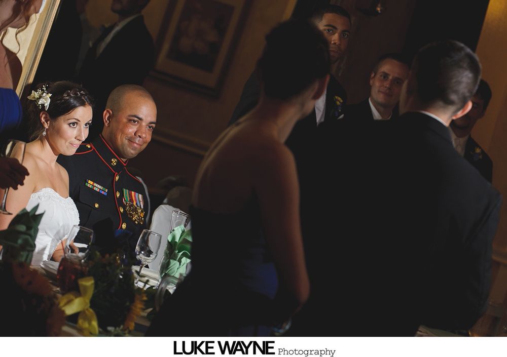 Bride and groom at a wedding reception; military uniform, formal attire, dark setting.