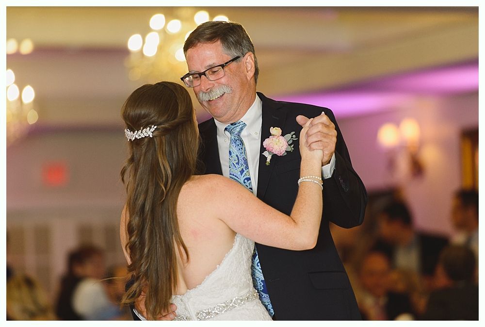 Bride and groom dancing at a wedding reception; she is laughing, wearing a white dress, and he wears a blue vest.