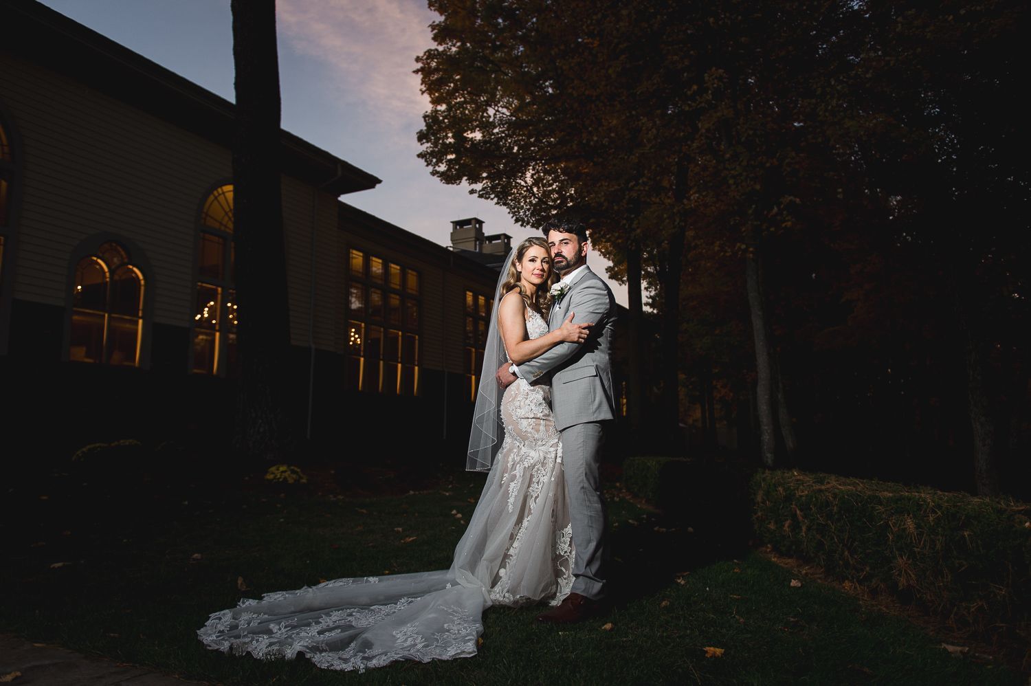 Bride and groom embrace outdoors at dusk, wedding dress with long train. Building with lit windows in background.