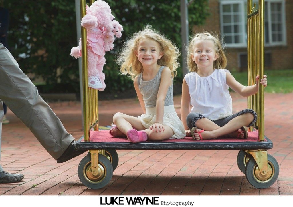 Two girls smiling, sitting on a luggage cart holding a pink stuffed animal. Outside, a person’s legs are pulling the cart.