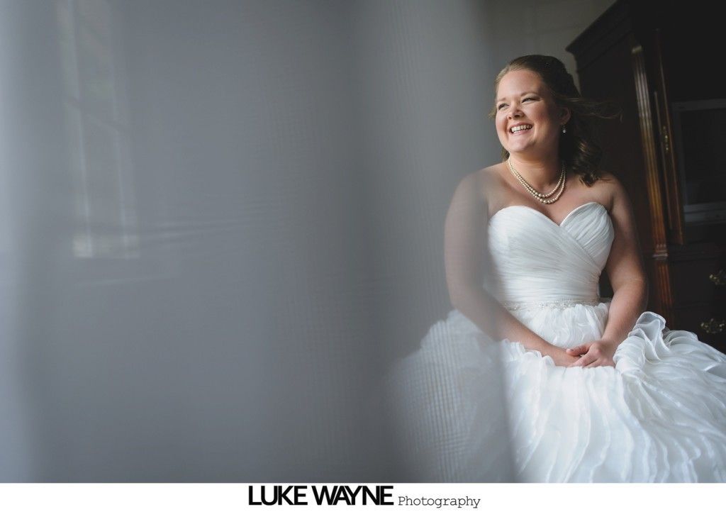 Bride in a white strapless gown smiles, seated near a window, with soft focus on the left.