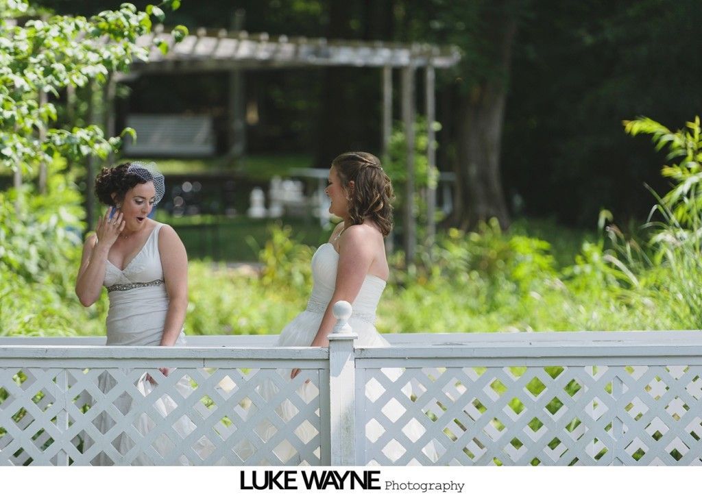 Two women in wedding dresses, talking outdoors by a white fence. One touches her face, smiling. Green foliage in background.