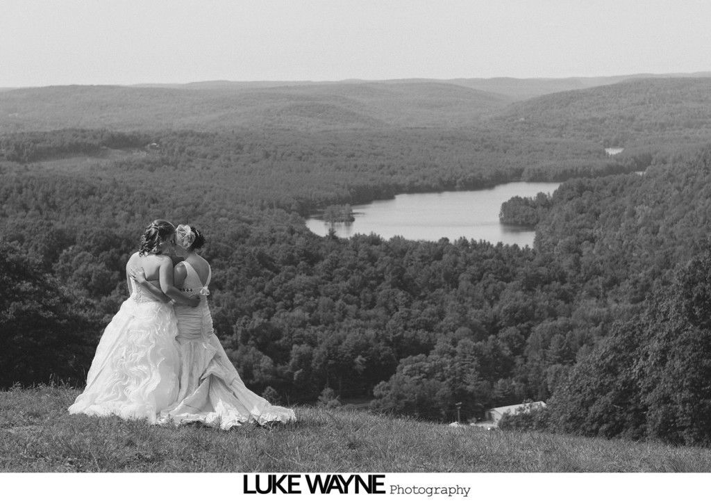 Two brides in wedding dresses embrace on a hill overlooking a lake and forest.
