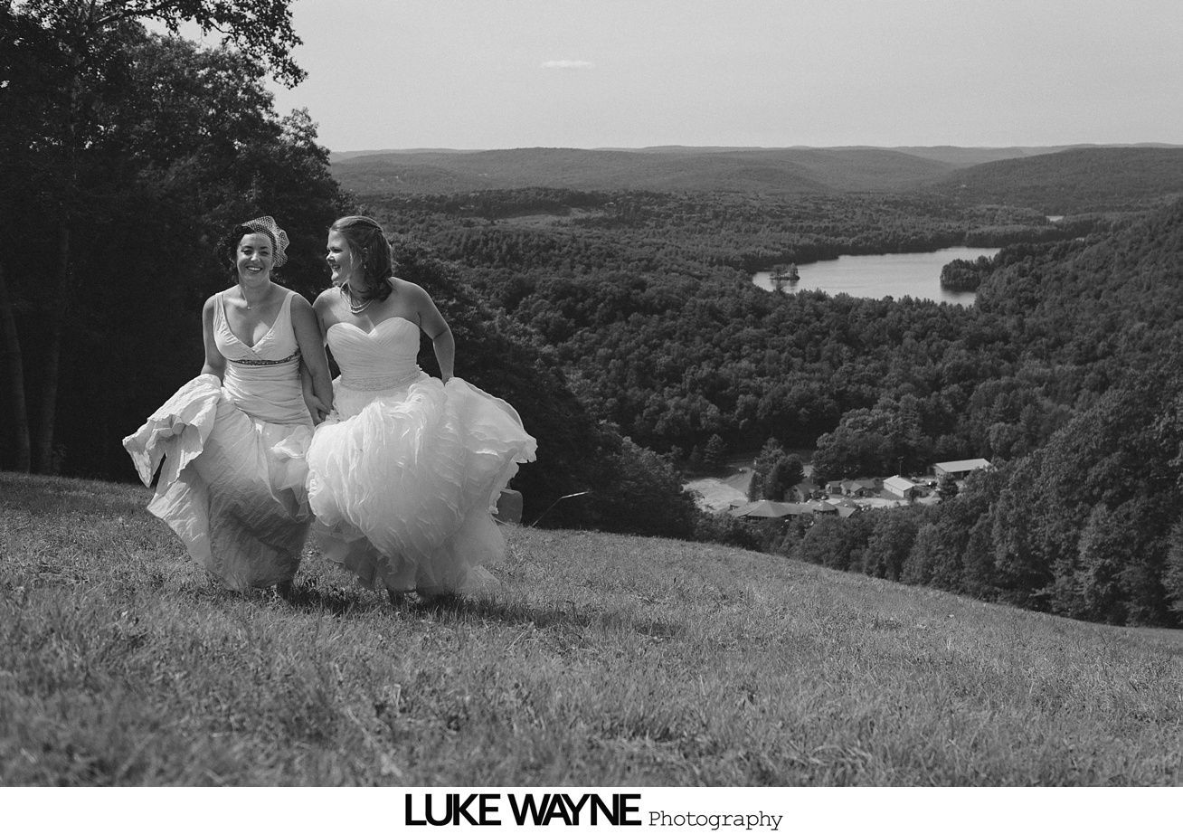 Two brides in wedding dresses walk hand-in-hand in a grassy field, overlooking a lake and forested hills.