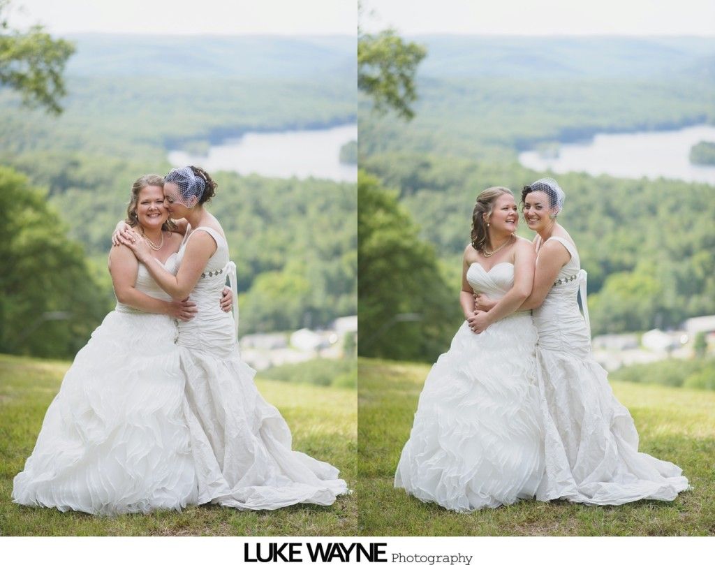 Two brides in white gowns embrace, smiling, against a scenic landscape with water and trees.