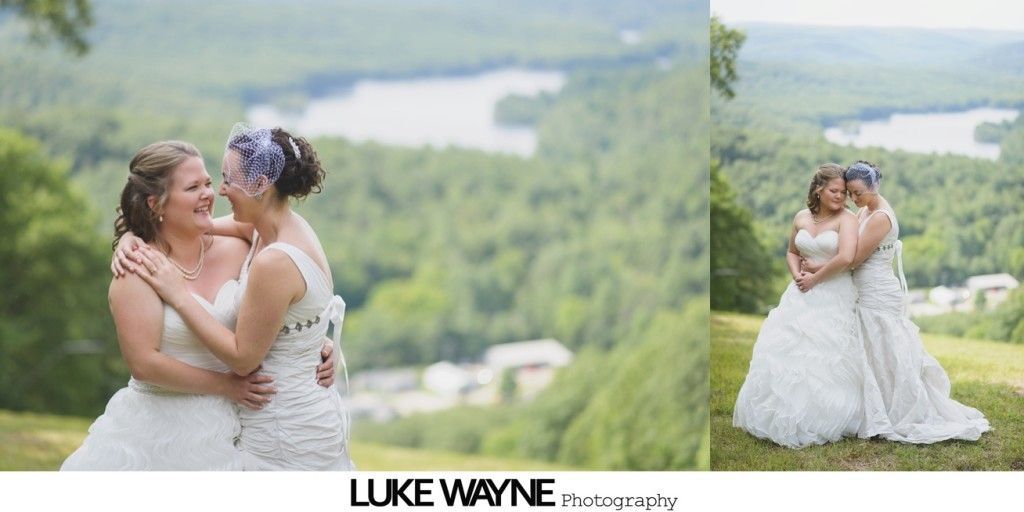 Two brides in white dresses embracing, laughing, with a scenic background of a lake and green trees.