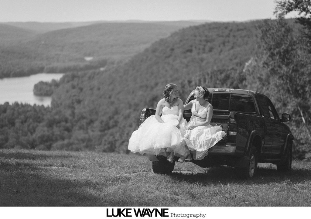 Two women in wedding dresses sit in the bed of a truck, overlooking a lake and hills.