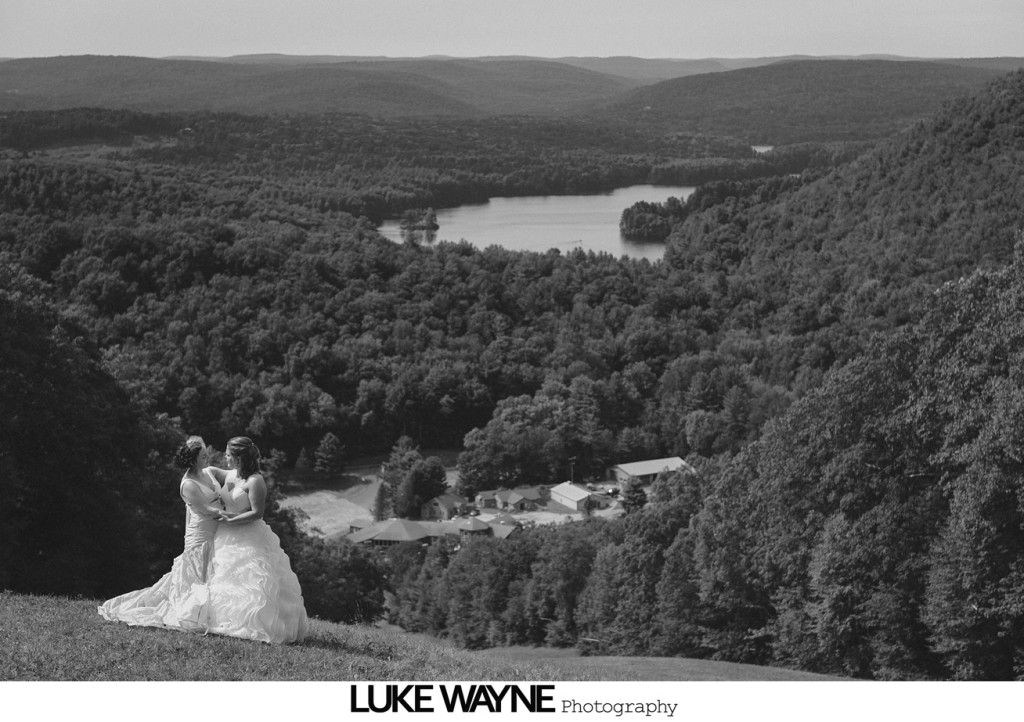 Two brides in wedding dresses embrace, overlooking a lake surrounded by forest.
