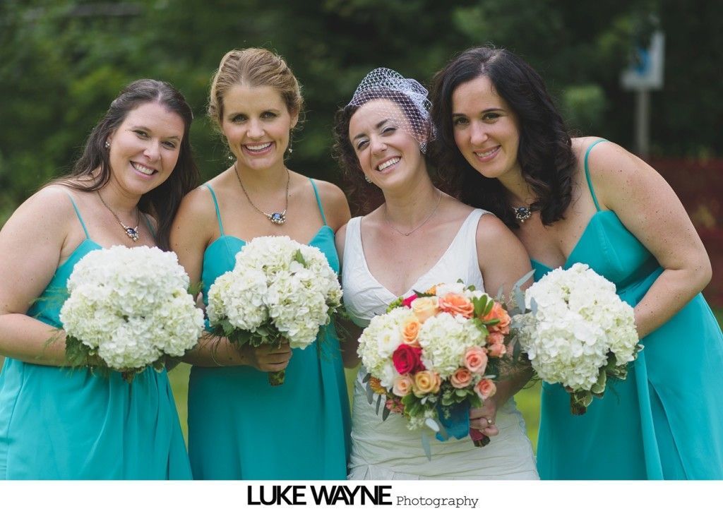 Bride with bridesmaids, posing outdoors. Bride holds bouquet; bridesmaids wear teal dresses, holding white flowers.