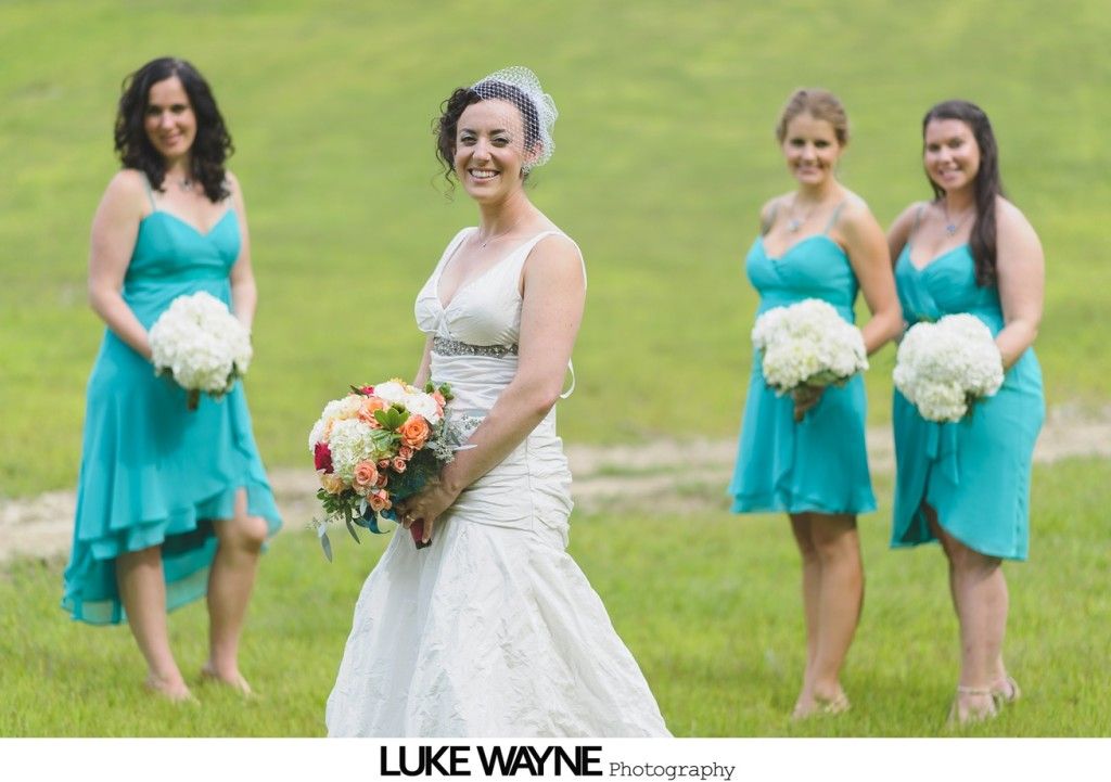Bride in white dress, bridesmaids in teal dresses, holding bouquets, on grassy field.