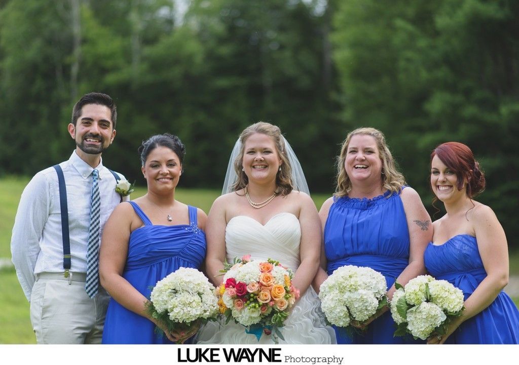 Wedding group in front of trees; bride in white gown, bridesmaids in blue dresses, and groom in blue tie.
