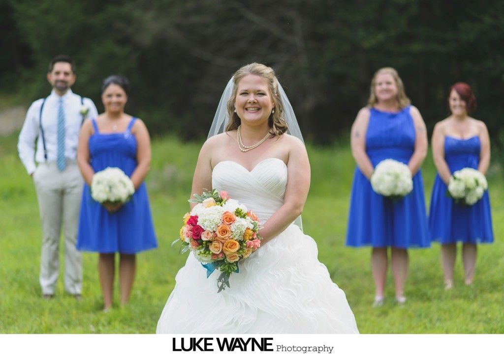 Bride smiling, holding flowers, with bridesmaids in blue dresses and a man in a field.