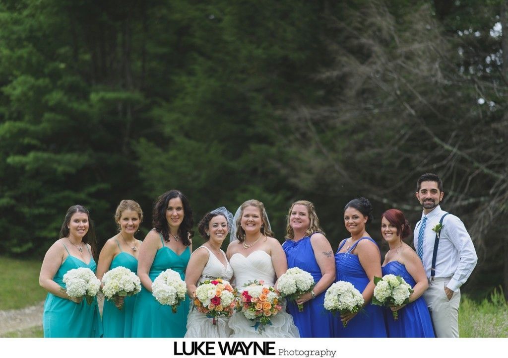 Bridesmaids in teal and blue dresses with bride in white gown, holding bouquets, and groomsman, posing outdoors.