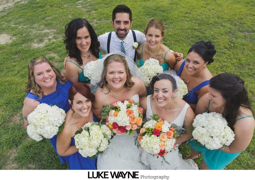 Wedding group poses in a field: bride, groom, bridesmaids smiling, holding flowers.