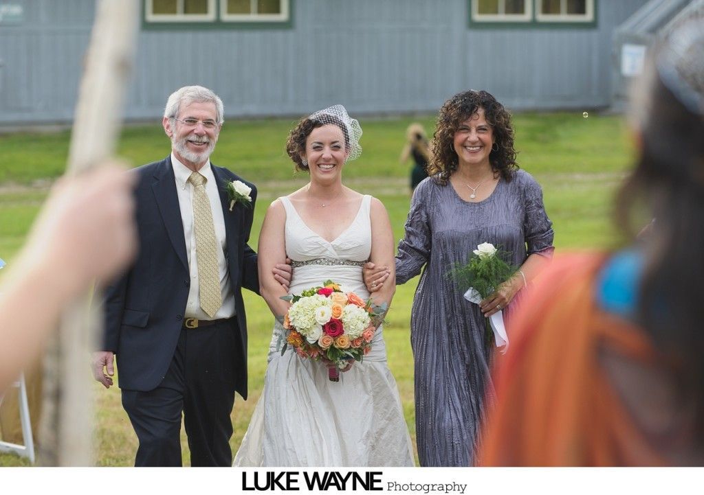 Bride escorted by parents, walking toward the camera at outdoor wedding; bride holding bouquet.