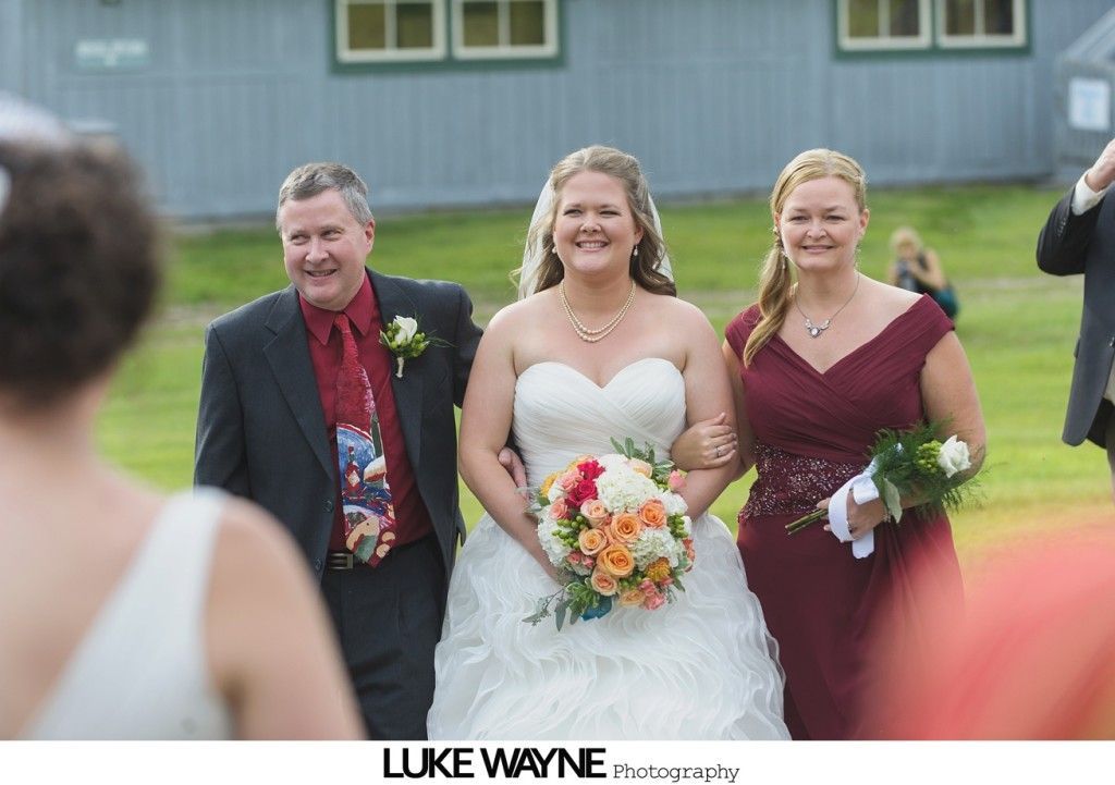 Bride escorted down aisle by her father and bridesmaid. The bride wears a white gown and holds a bouquet.