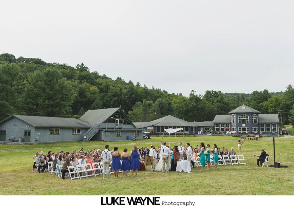 Wedding ceremony in a field with guests facing a couple. Buildings and trees in the background.