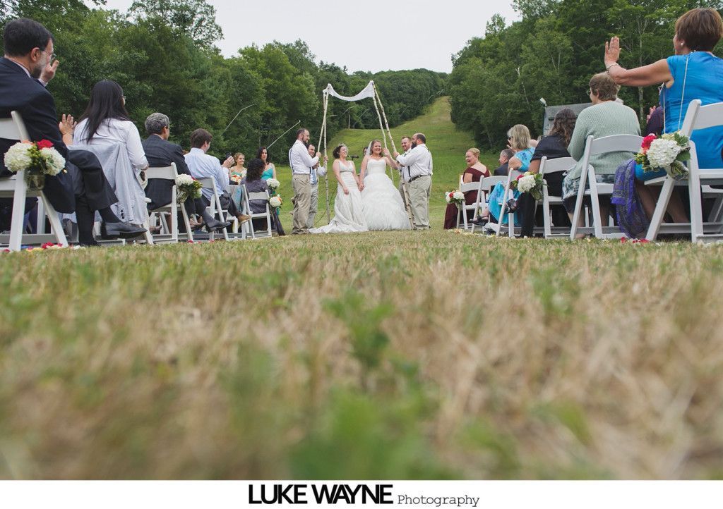 Wedding ceremony with two brides under a decorated arch, guests seated on white chairs in a grassy area.