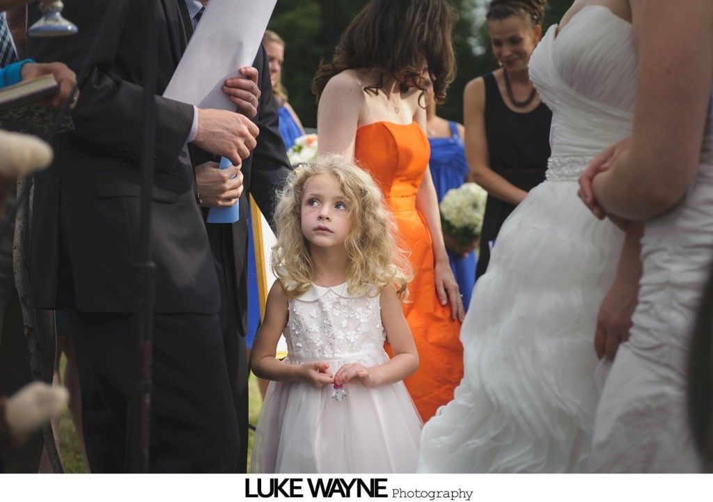 Flower girl watches wedding ceremony; bride in white gown, bridesmaid in orange dress.
