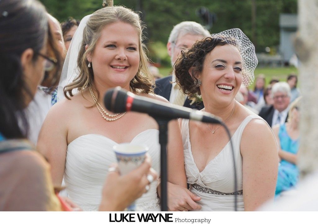 Two women in white dresses smiling during a wedding ceremony outdoors, with a microphone in front of them.