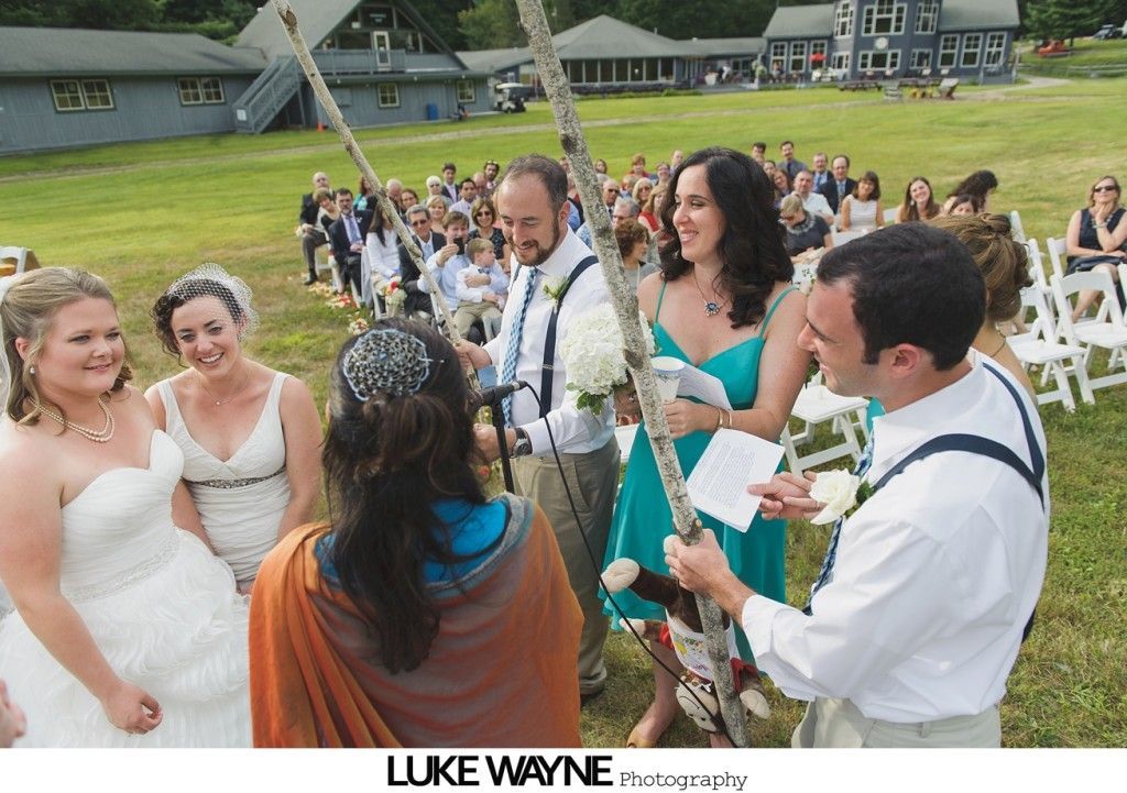 Wedding ceremony outdoors; two brides, officiant, guests, and a celebrant with flowers and white birch arch.