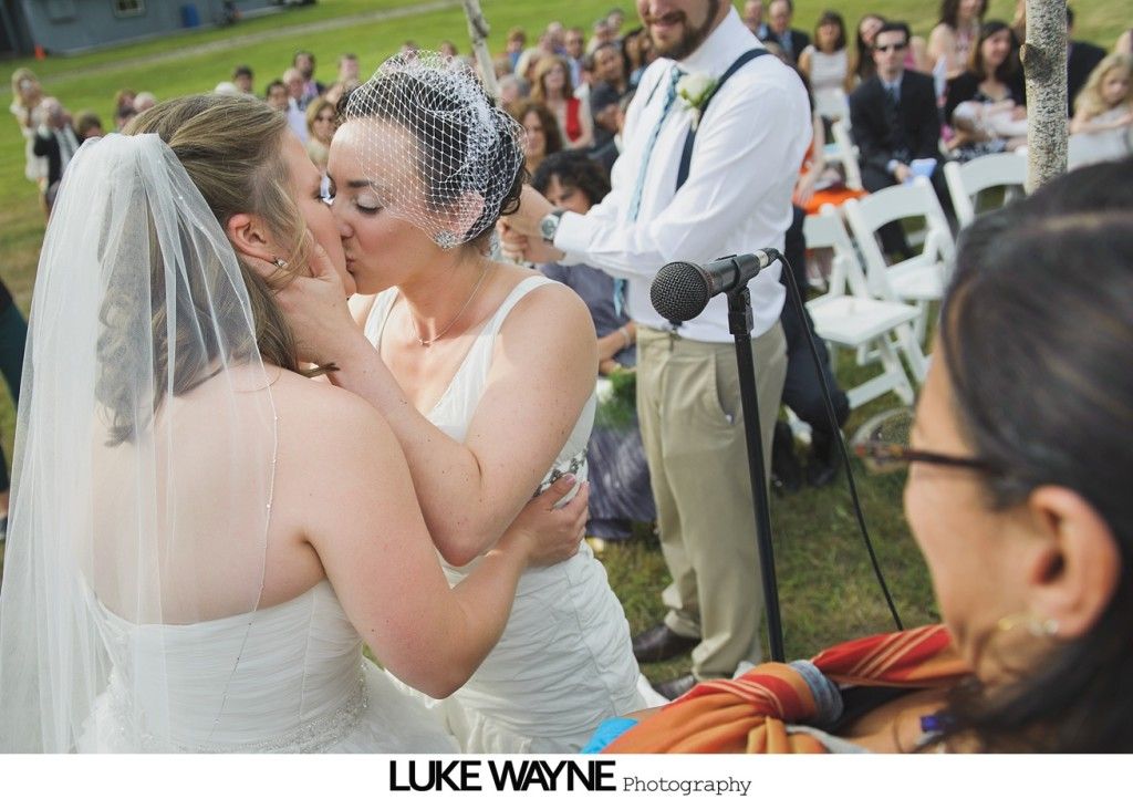 Two brides kiss at an outdoor wedding ceremony; guests and officiant watch.
