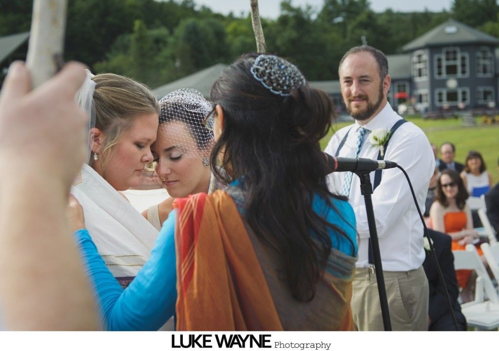 Wedding ceremony: two brides in white and blue embrace, officiant in orange, man with microphone nearby, outdoor setting.