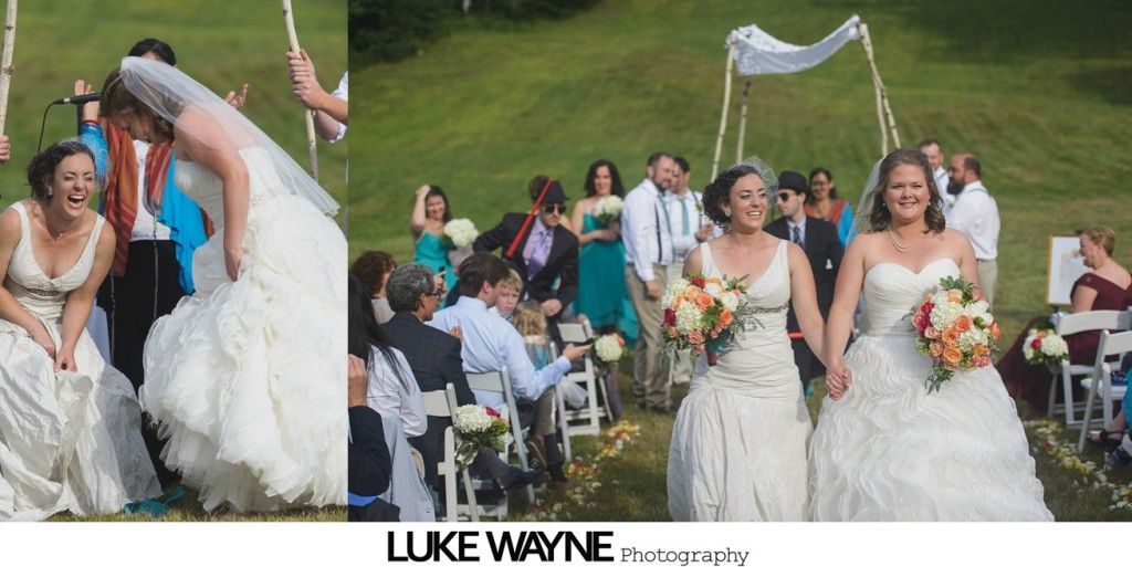 Two brides in white gowns hold hands after an outdoor wedding ceremony. Guests and wedding party in background.