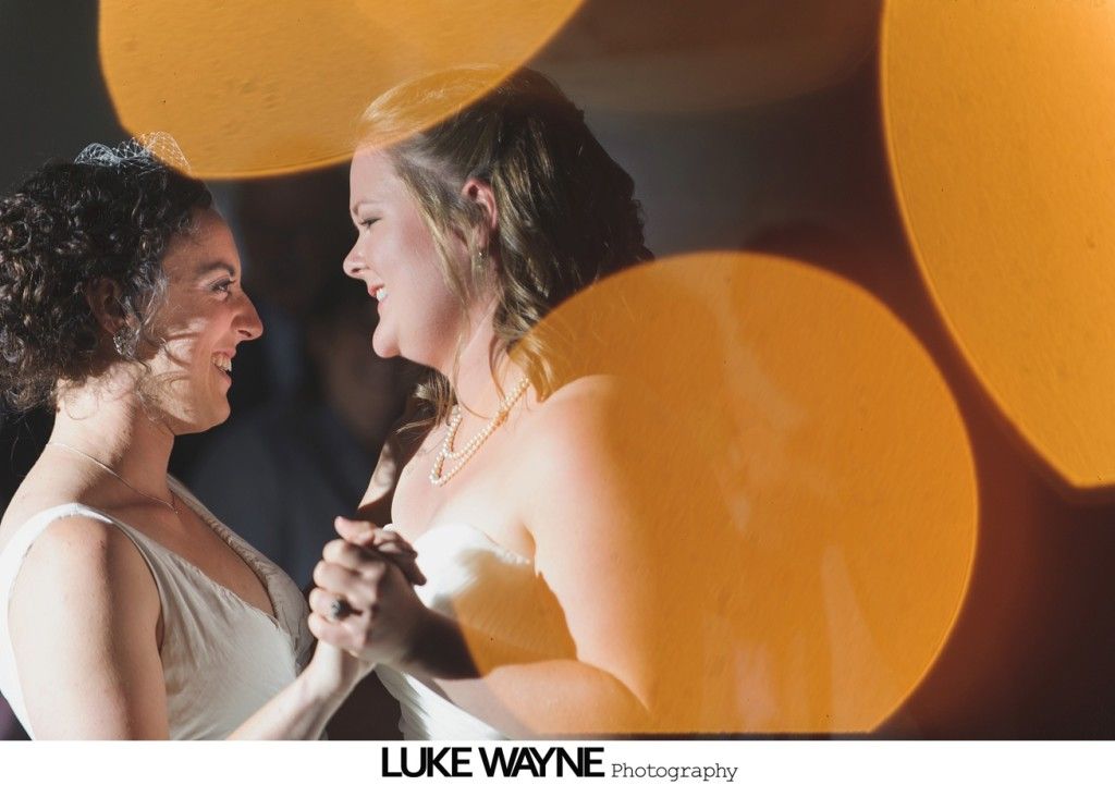 Two women in white dresses dancing, smiling, lit by warm yellow bokeh.