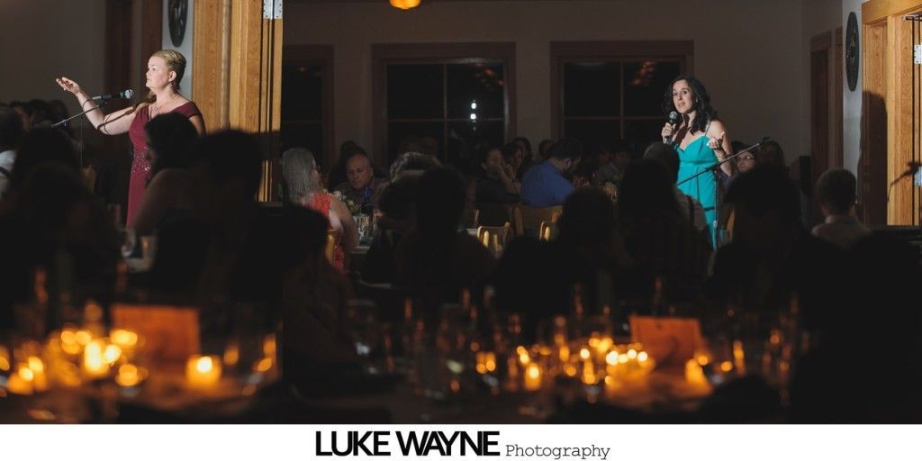 Two women singing at an event with candles and people. One in red dress, one in blue dress.