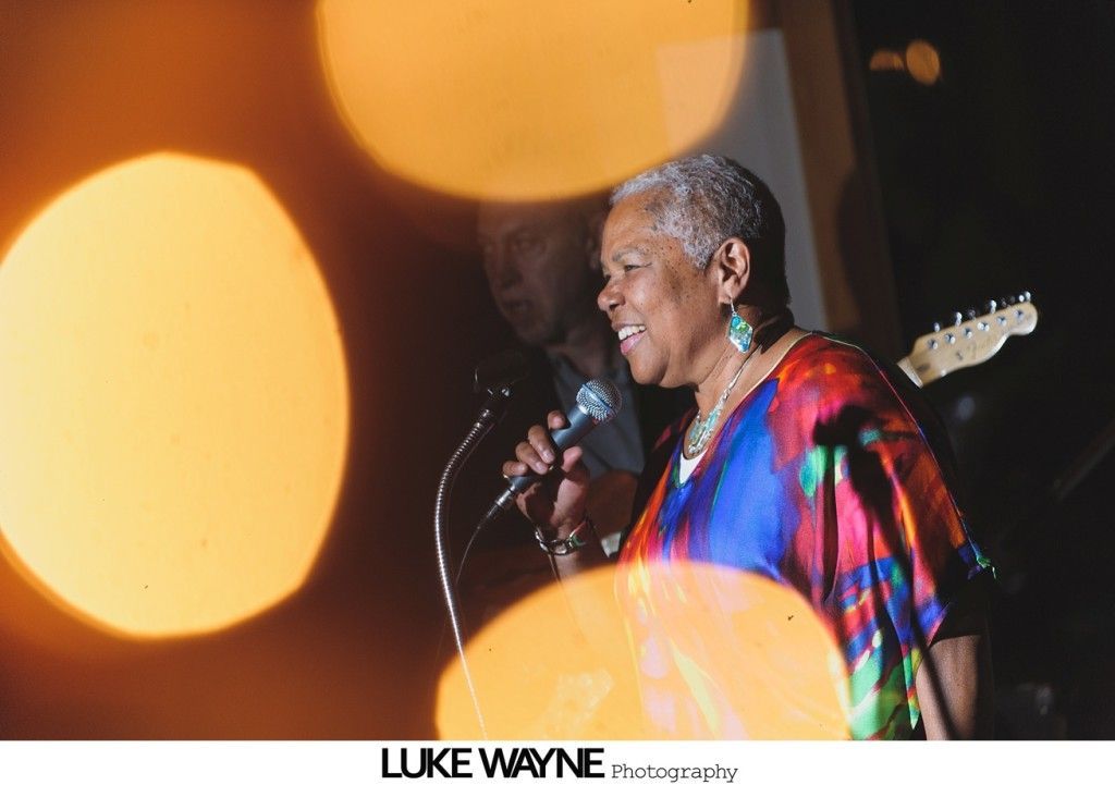 Woman singing into a microphone onstage; blurry orange lights in the foreground.