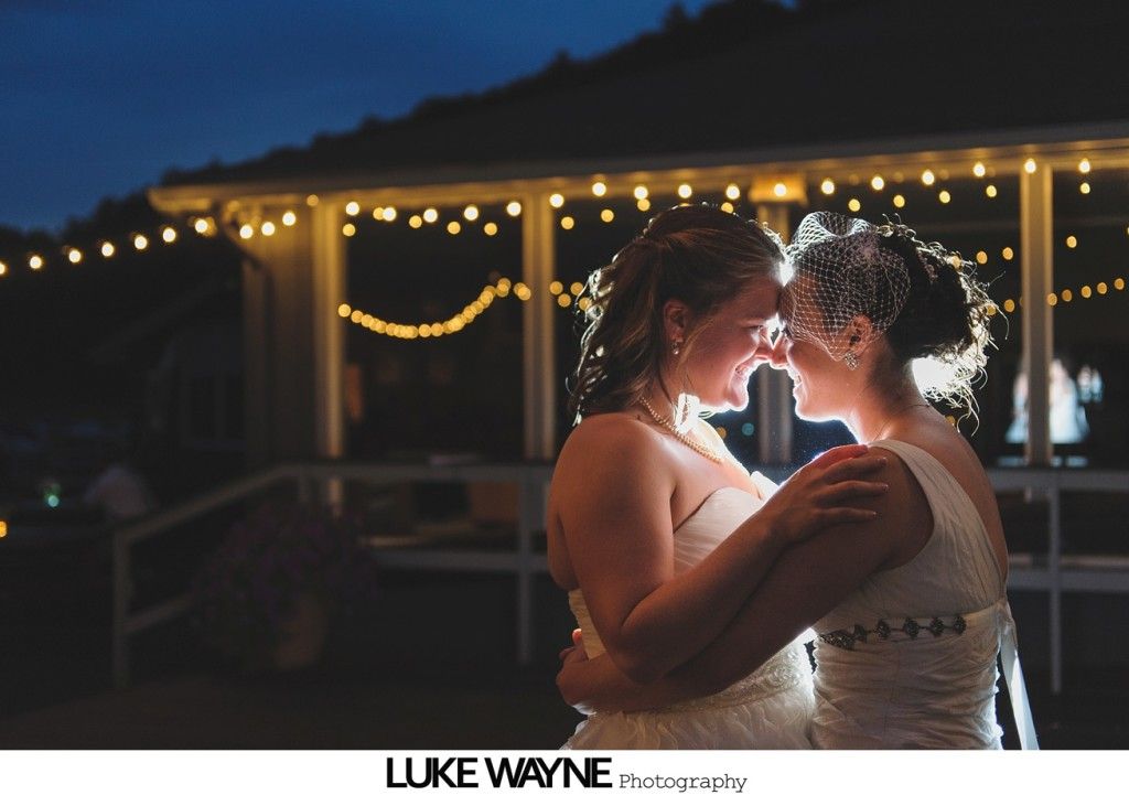 Two brides embrace, illuminated by string lights, at night.