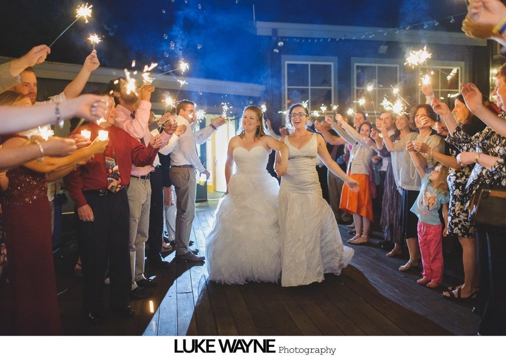 Two brides walk through a sparkler-lit exit, surrounded by cheering guests on a wooden deck.
