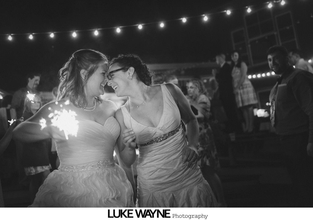 Two brides laughing, holding sparklers at an outdoor wedding reception. String lights in the background.