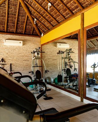 Academia com aparelhos de ginástica, espelhos e telhado de palha.