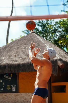 Homem de sunga jogando bola na praia. Chapéu de sol, bola marrom, cabana de palha ao fundo.