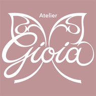 Logo bianco “Atelier Gioia” su sfondo rosa cipria, stilizzato come ali di farfalla e con scritta in corsivo.
