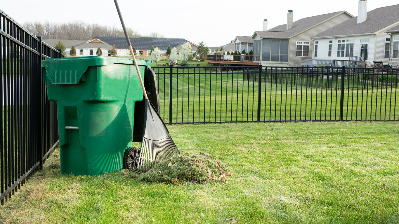 Green trash bin beside a black fence in a grassy suburban yard with houses in the background