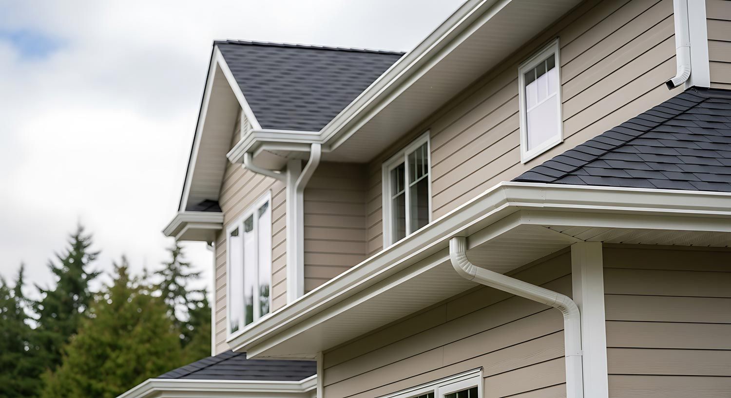 Close-up of a beige house exterior with white trim, gray shingle roof, and evergreen trees behind it Close-up of a beige house exterior with white trim, gray shingle roof, and evergreen trees behind it