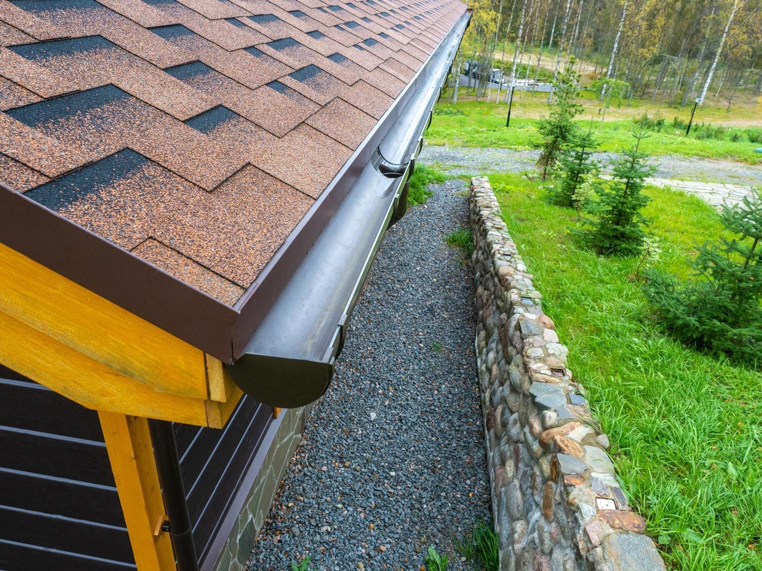 Close-up of a house roof and gutter beside a gravel path and stone retaining wall in a landscaped yard Close-up of a house roof and gutter beside a gravel path and stone retaining wall in a landscaped yard