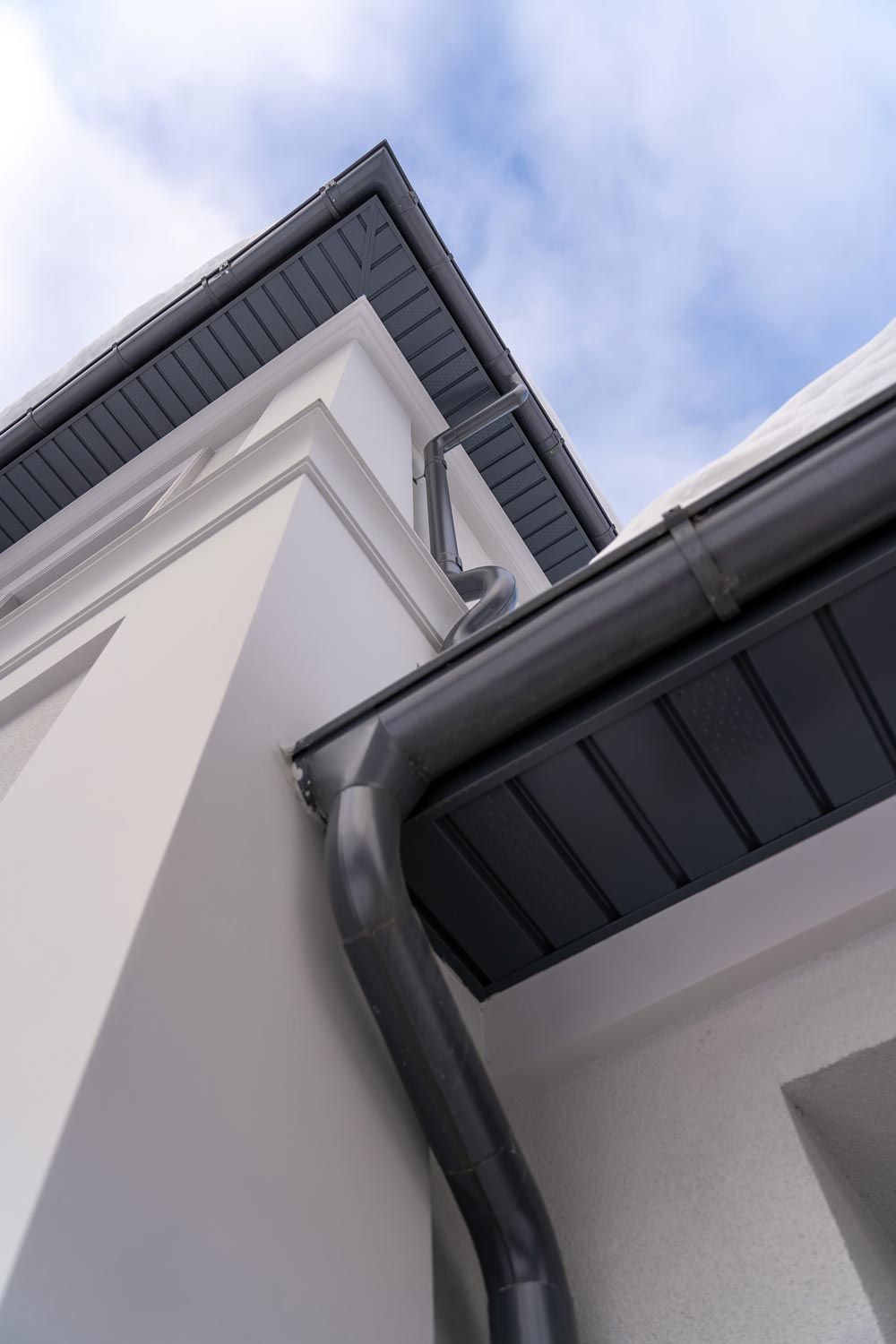 Upward view of a white house exterior with dark gray rooflines and a gutter against a cloudy sky Upward view of a white house exterior with dark gray rooflines and a gutter against a cloudy sky