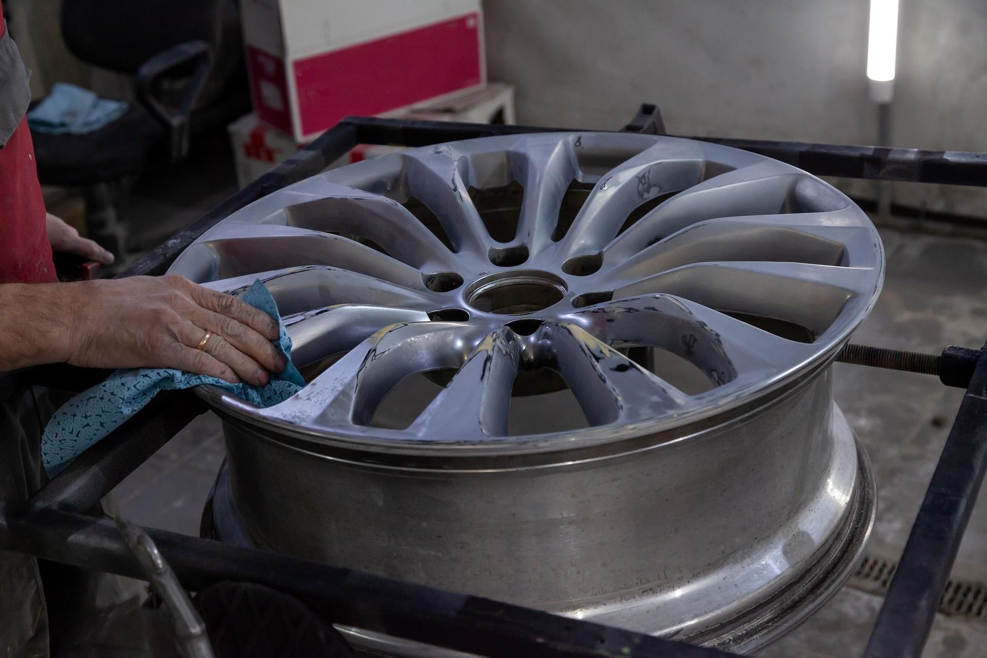 Person cleaning a silver car wheel with a blue cloth; the wheel is on a black stand indoors.