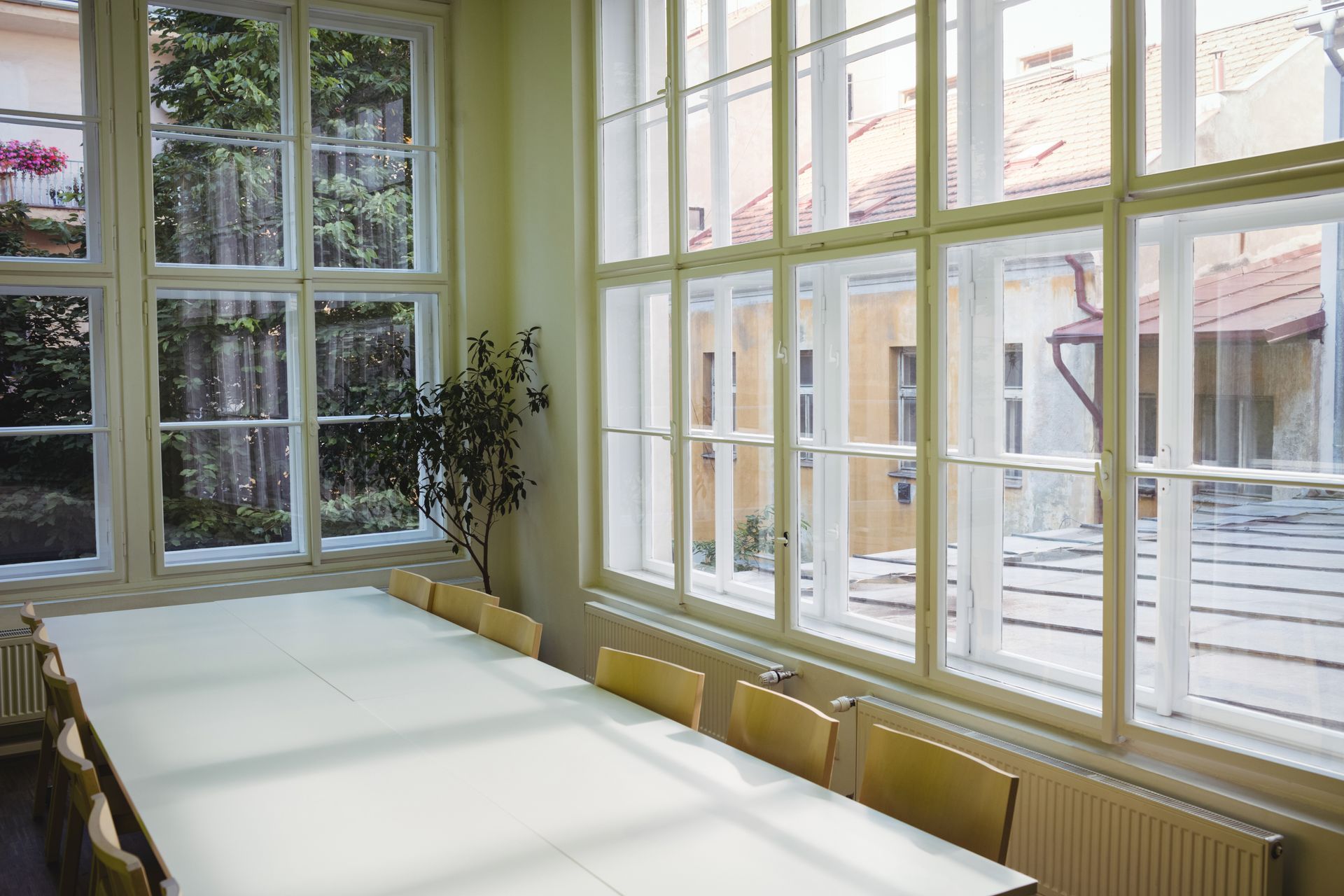 Bright sunlit room with large windows, white tables, and yellow chairs overlooking an outdoor courtyard